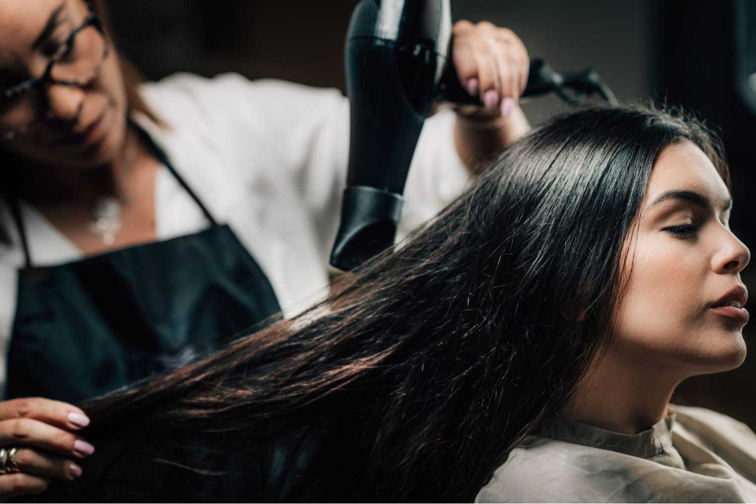 Hair stylist blow-drying a woman's long hair in a salon setting.